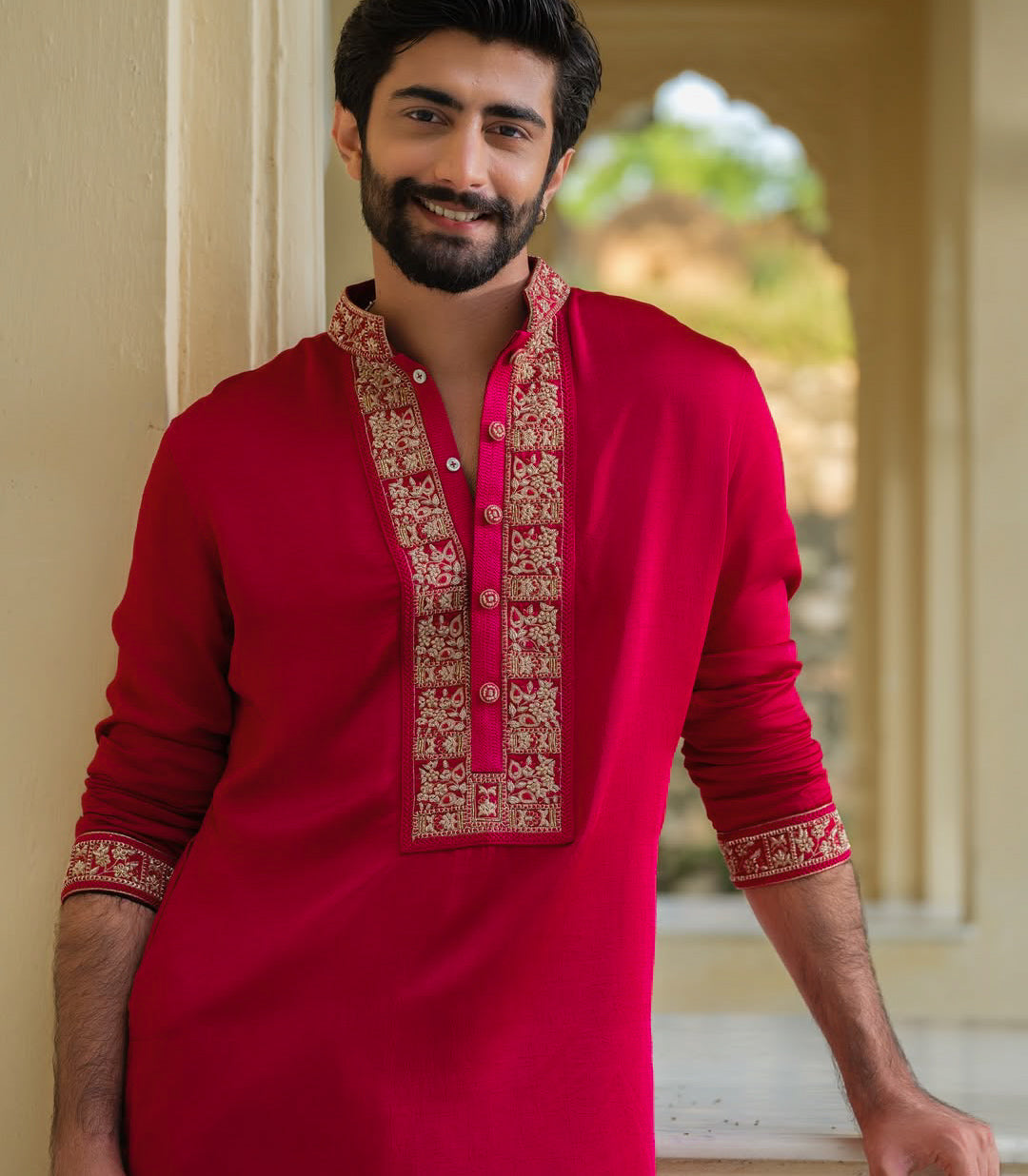 A smiling man wearing a bright red long-sleeve kurta with ornate gold embroidery on the front placket and cuffs, standing in a softly lit architectural setting.