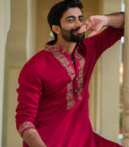 A man wearing a bright red long-sleeve kurta with ornate gold embroidery on the collar and cuffs, posing in a warm architectural setting.