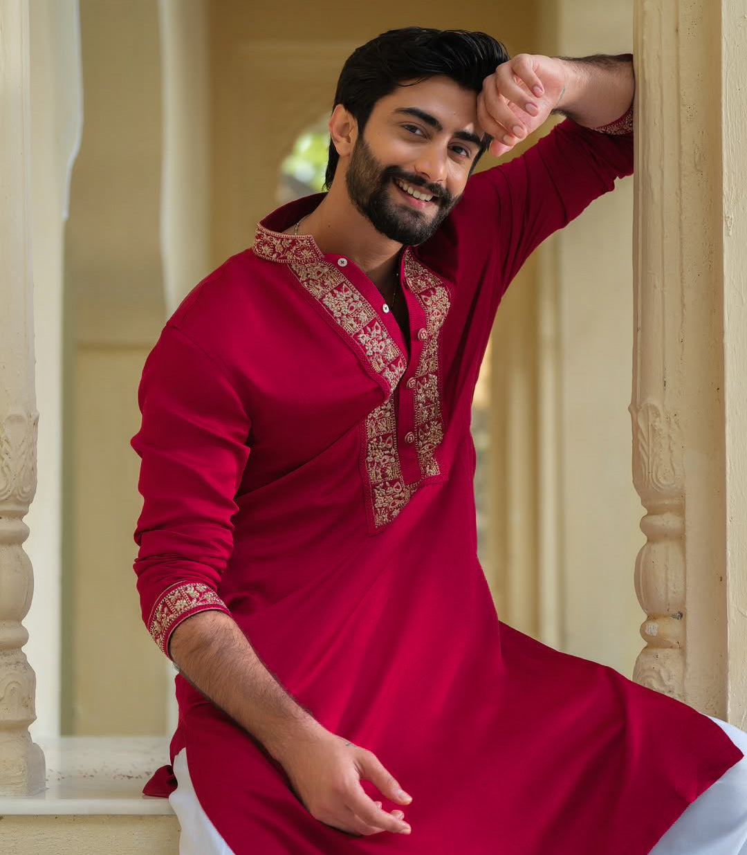 A man wearing a bright red long-sleeve kurta with ornate gold embroidery on the collar and cuffs, posing in a traditional architectural setting with cream-colored pillars.