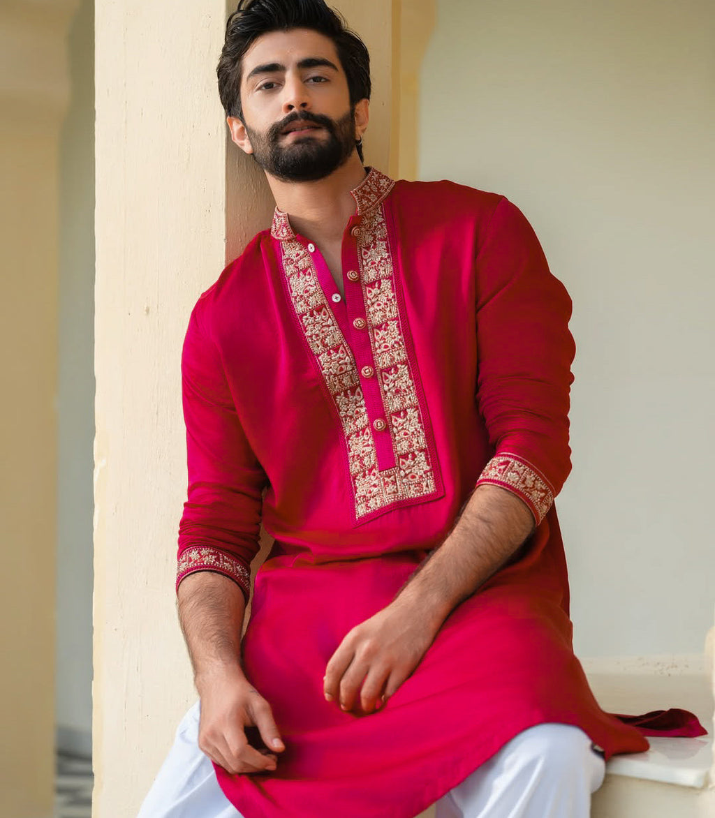 A man wearing a bright pink traditional kurta with ornate gold embroidery on the chest and cuffs, paired with white trousers, sitting in a neutral indoor setting.